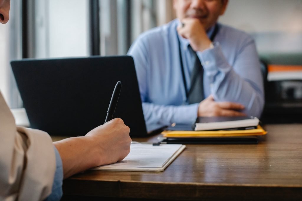 Employee writing with pen on pad of paper while supervisor sits across table.